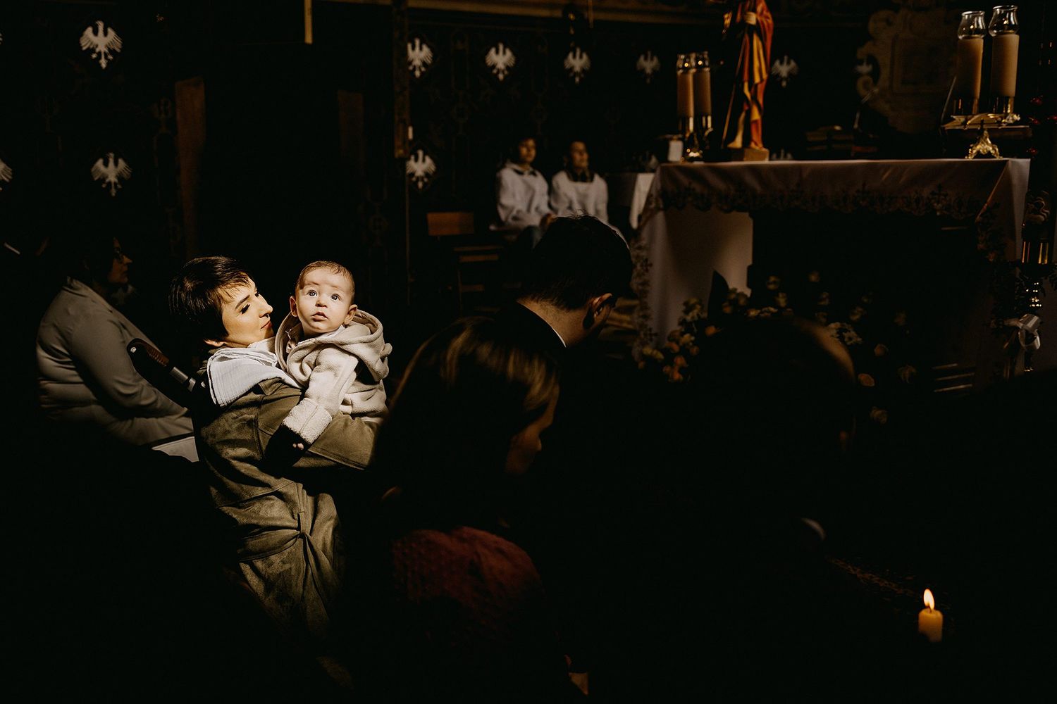 Person holds two babies near candlelit altar during intimate church baptism ceremony.