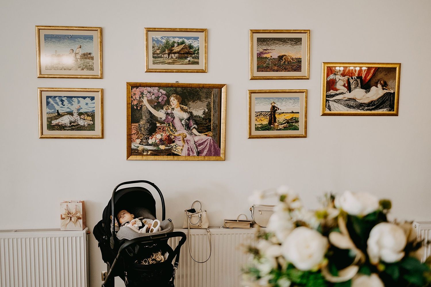 Baby sleeping in a black pram beneath a gallery wall of gold-framed paintings in a decorated room.