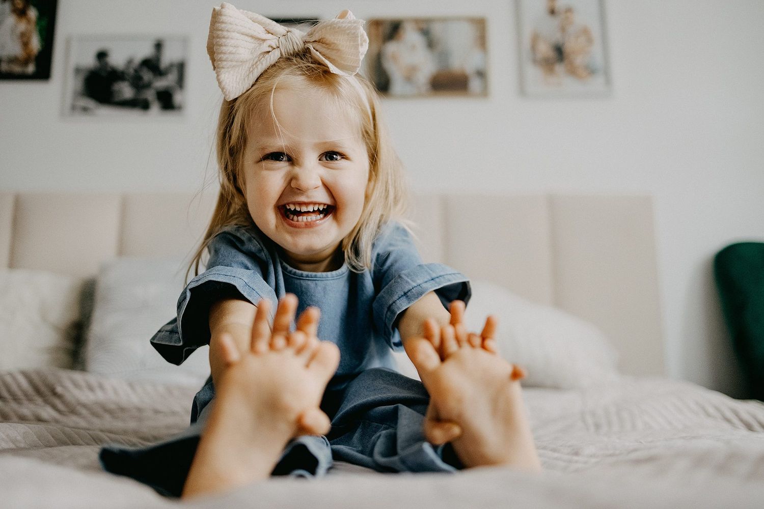 Adorable laughing toddler girl with a bow in her hair sitting on a white bed showing her feet.