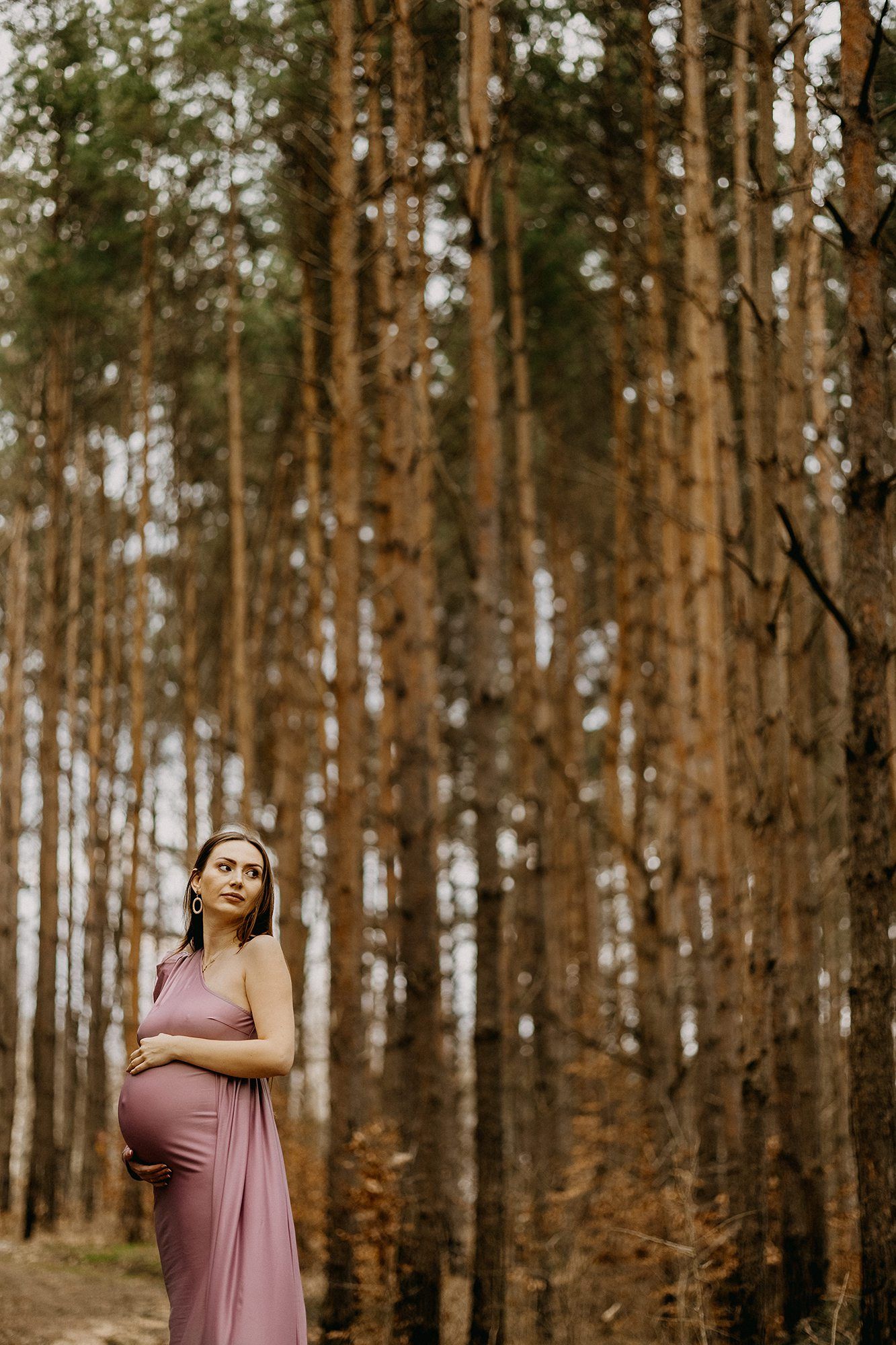 Pregnant woman in pink maxi dress standing among tall pine trees in autumn forest.