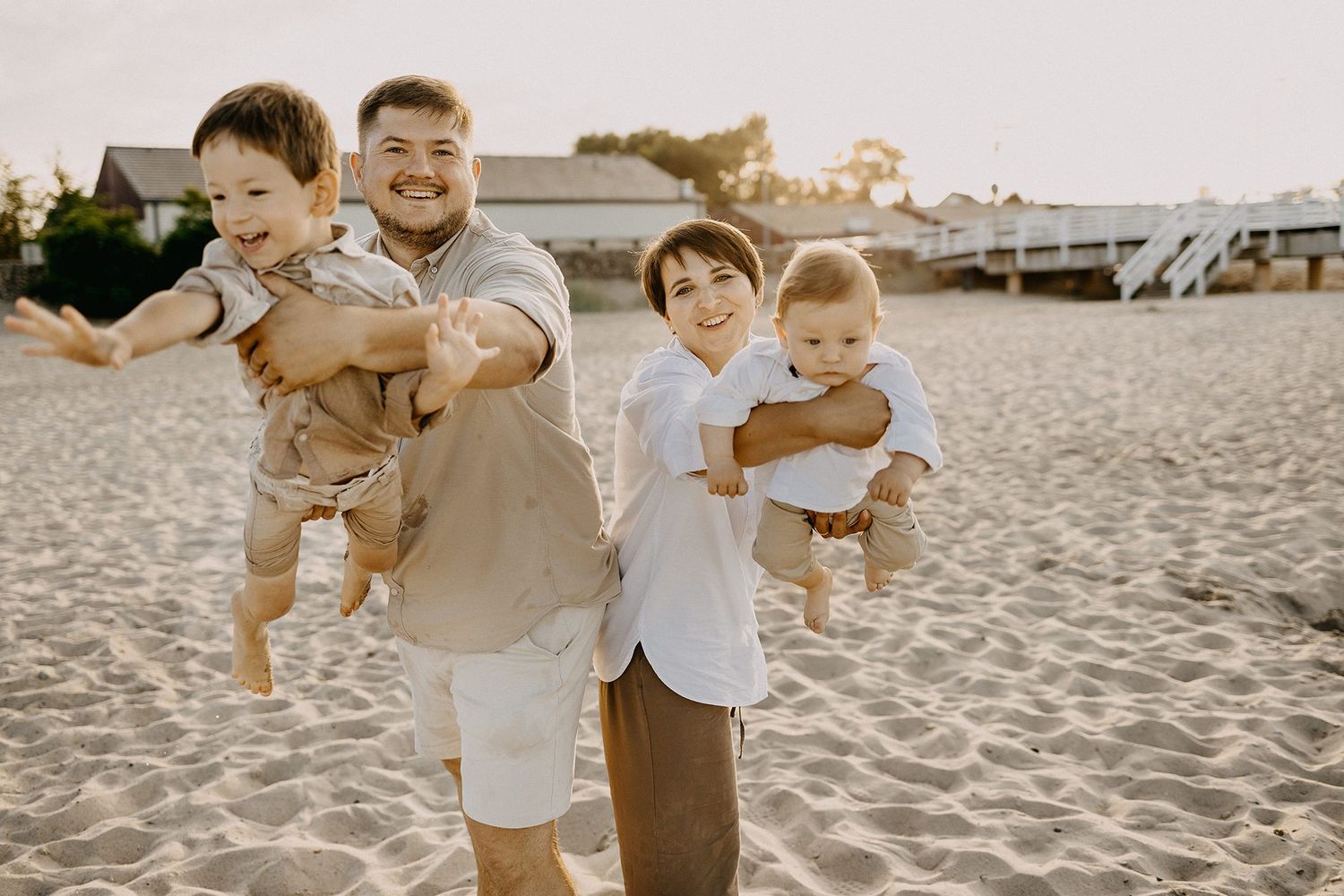 Smiling parents playfully holding two babies like airplanes on a sandy beach at sunset.