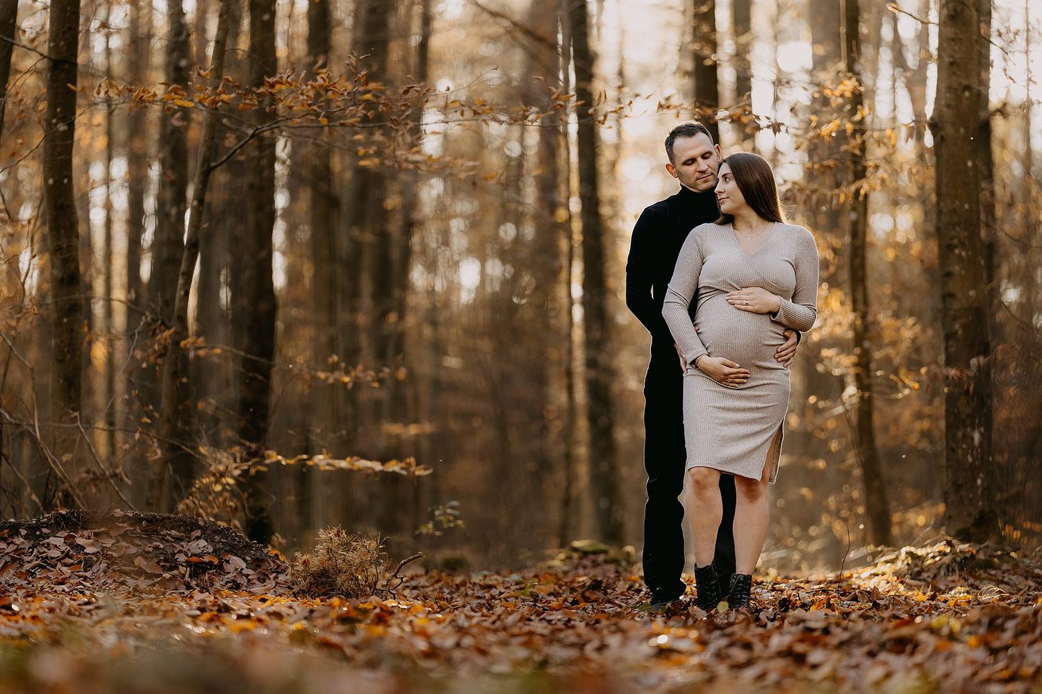 Couple embracing in autumn forest with man kissing pregnant womans head among fallen leaves.