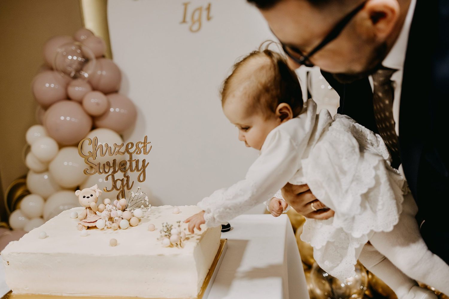 Baby in white christening dress reaches toward baptism cake with teddy bear cake topper.