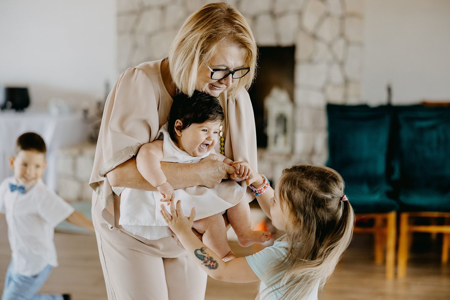 Blonde woman in beige outfit playfully holding baby with children around her indoors.