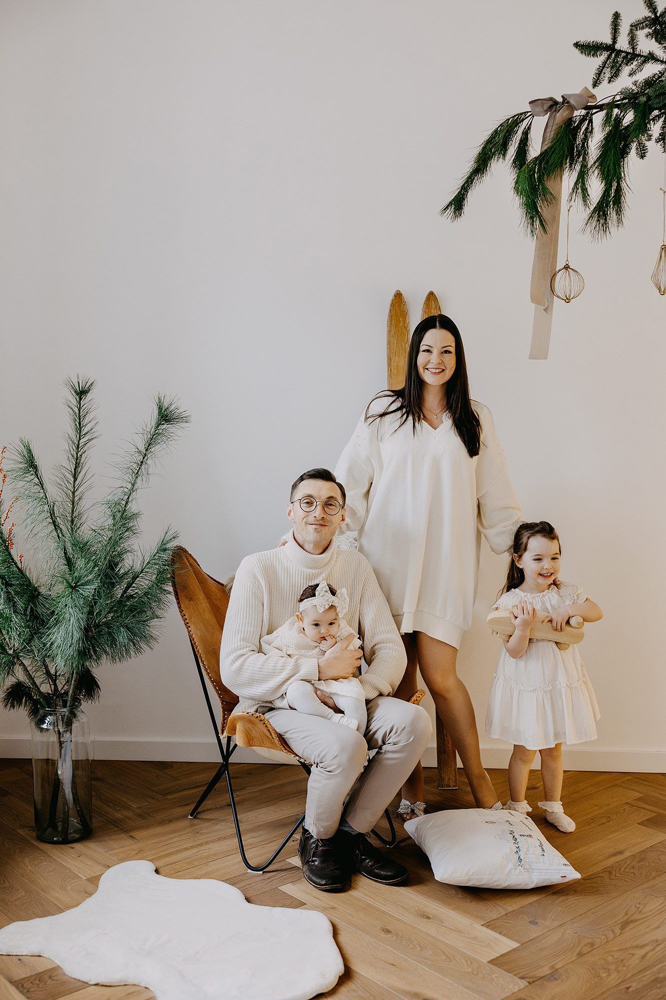 Young family with two small children posing in a minimalist white Christmas decorated room.