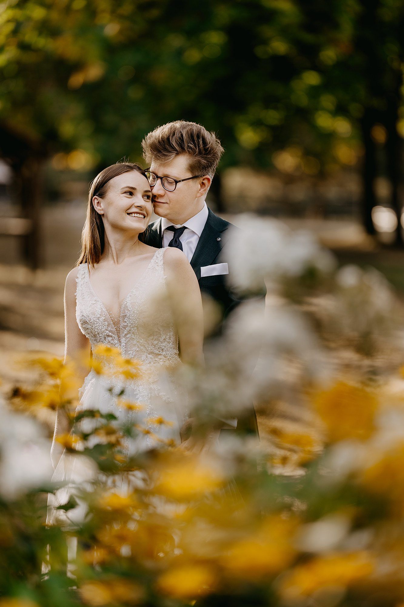 Smiling bride and groom with glasses posing among yellow and white flowers in autumn park.