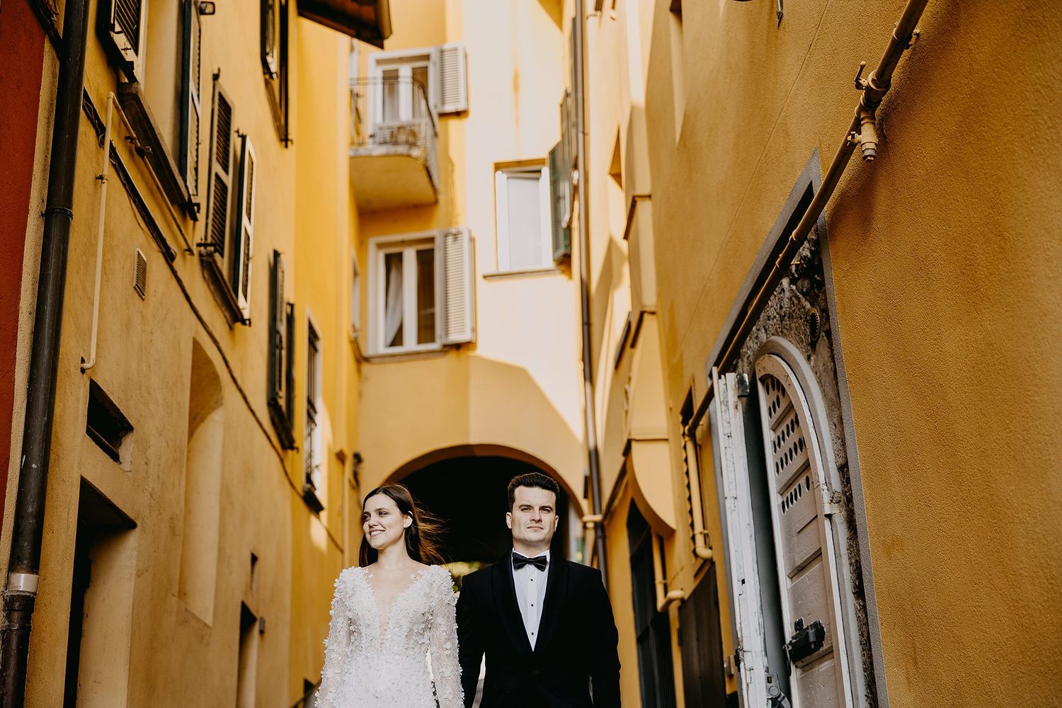 Bride and groom walk hand in hand through narrow Italian street lined with yellow buildings.