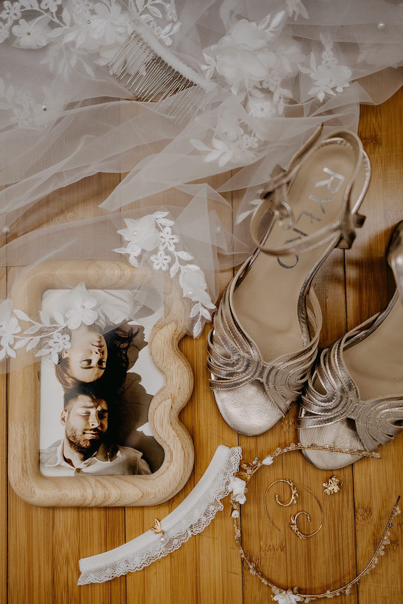 Bridal flat lay with lace veil, strappy heels, garter, jewelry, and a framed couple photo on wood.