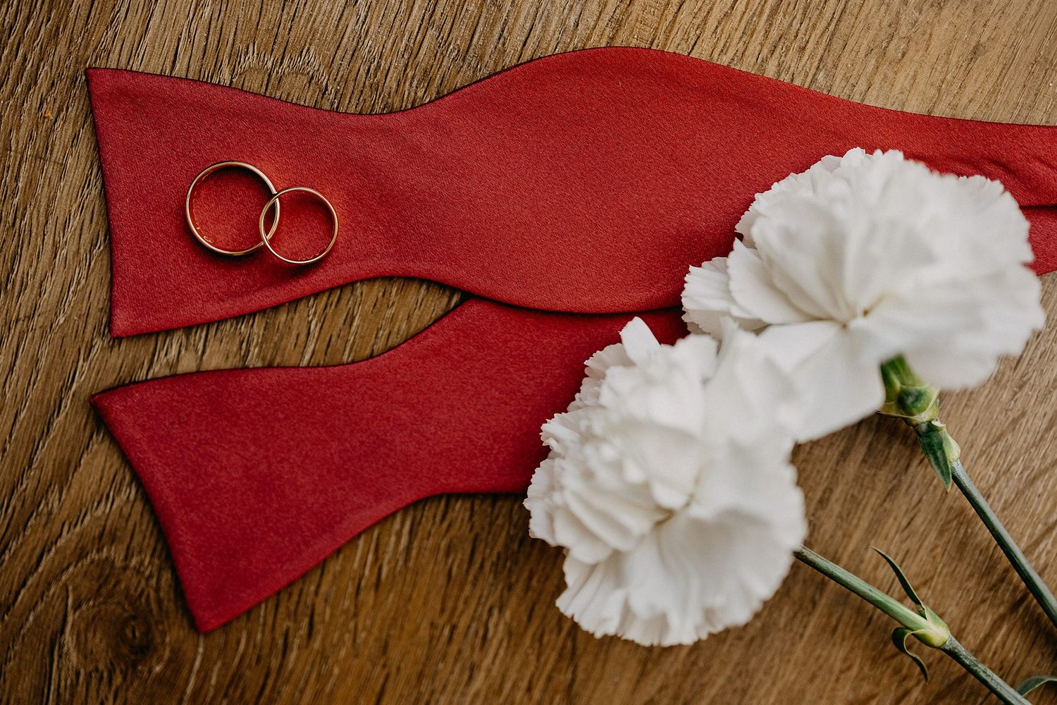 Two gold wedding rings resting on a red bow tie beside white carnation flowers.