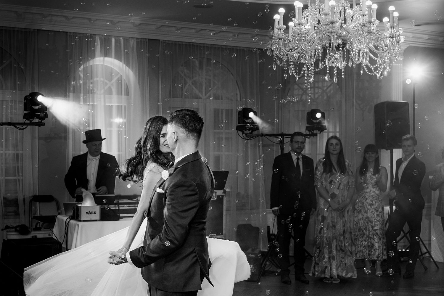 Bride and groom share first dance under a crystal chandelier in black and white wedding photo.