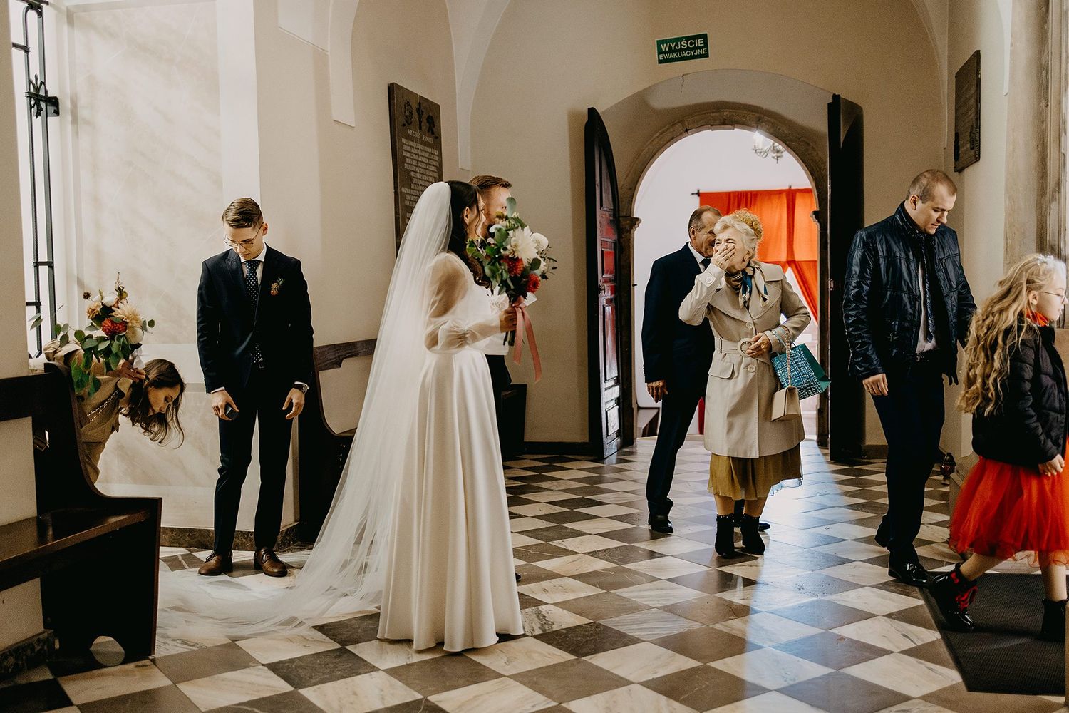 Bride in white gown holds bouquet while wedding guests gather in a historic church hallway with checkered floors.