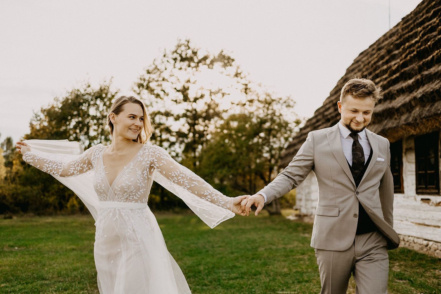 Bride in lace gown and groom in gray suit holding hands near a rustic thatched-roof building.