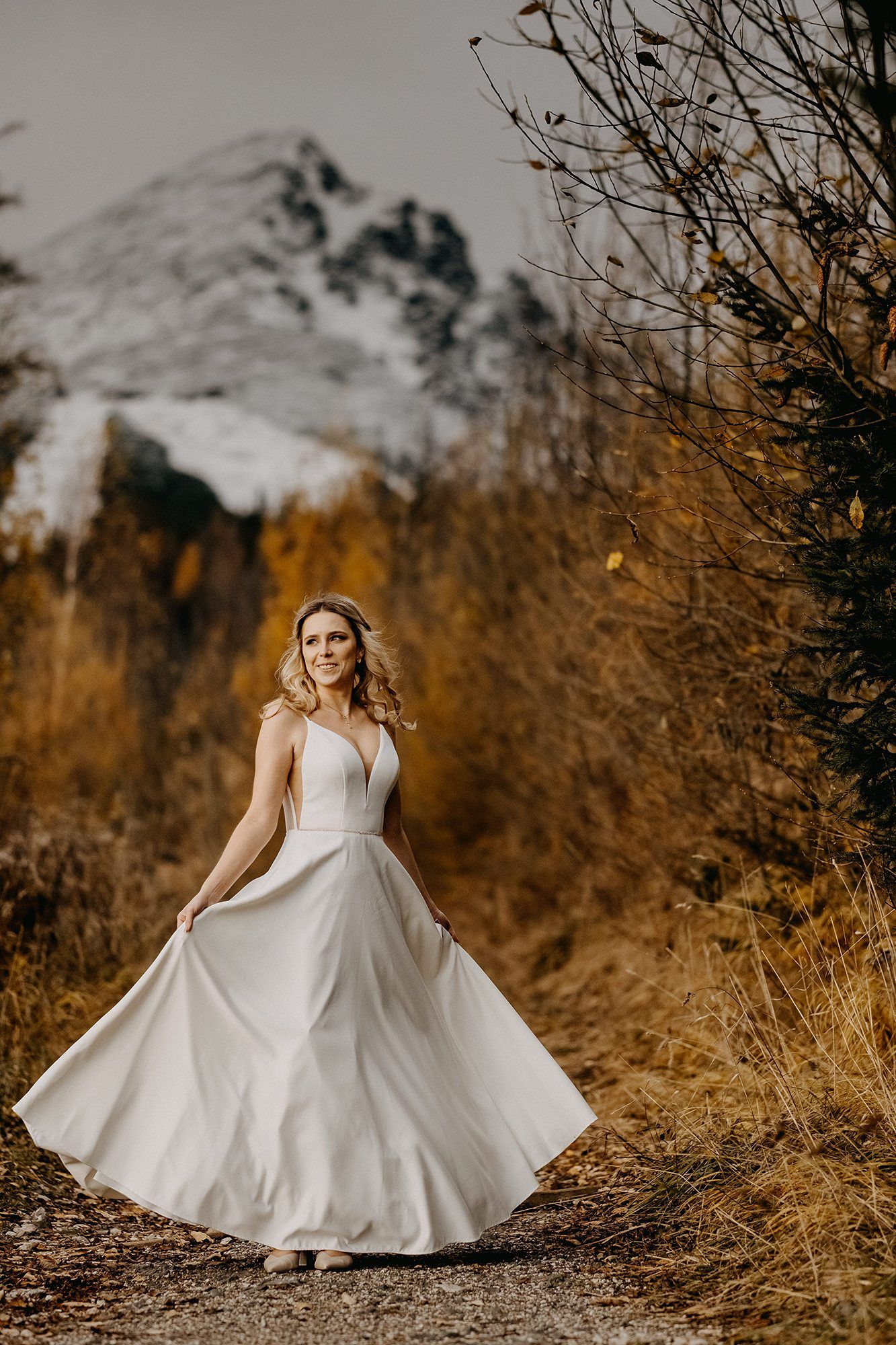 Bride in white gown twirls on a path surrounded by golden foliage and snowy mountains.
