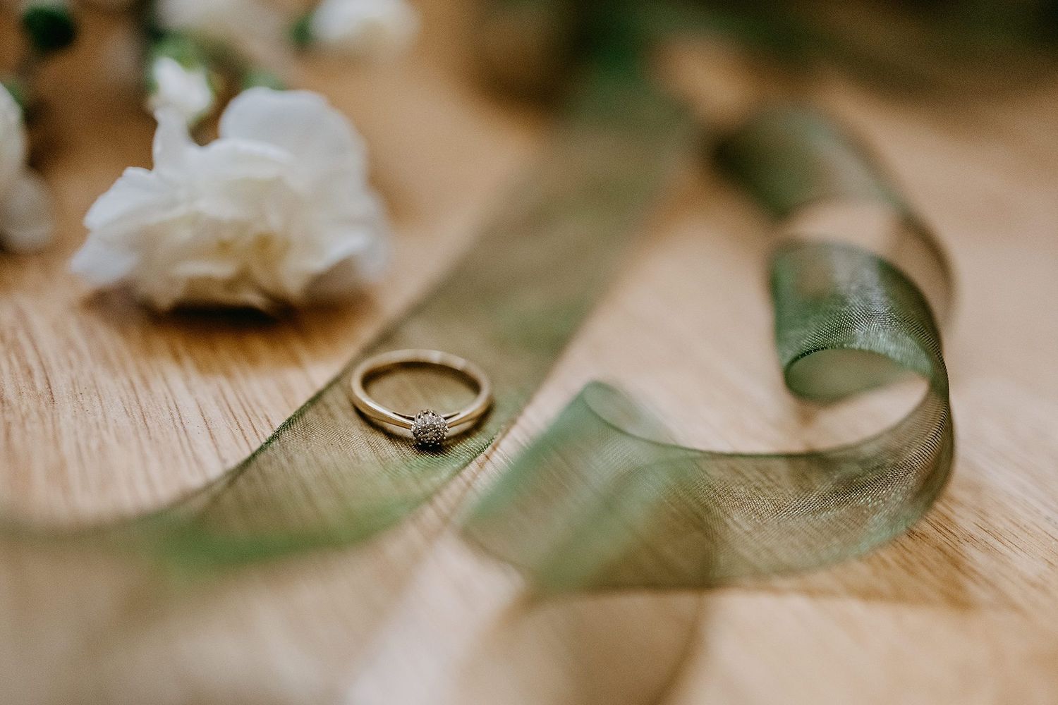 Gold diamond engagement ring resting on a green satin ribbon with white flowers on a wooden surface.