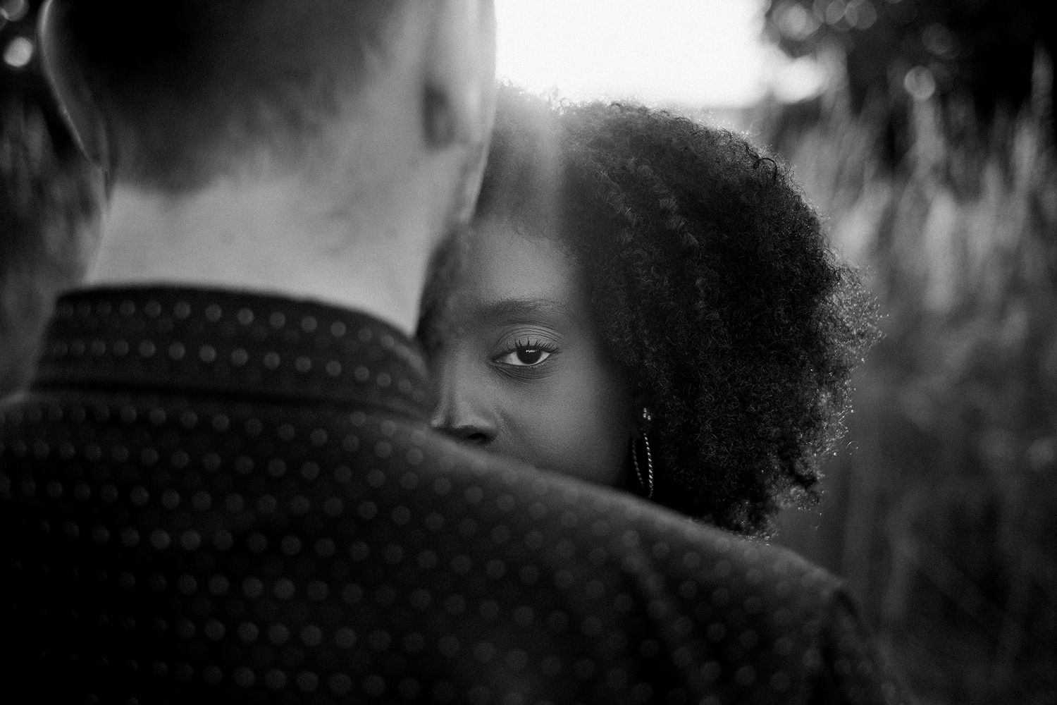 Black and white portrait of a woman with natural hair peering over a man's shoulder in soft backlight.