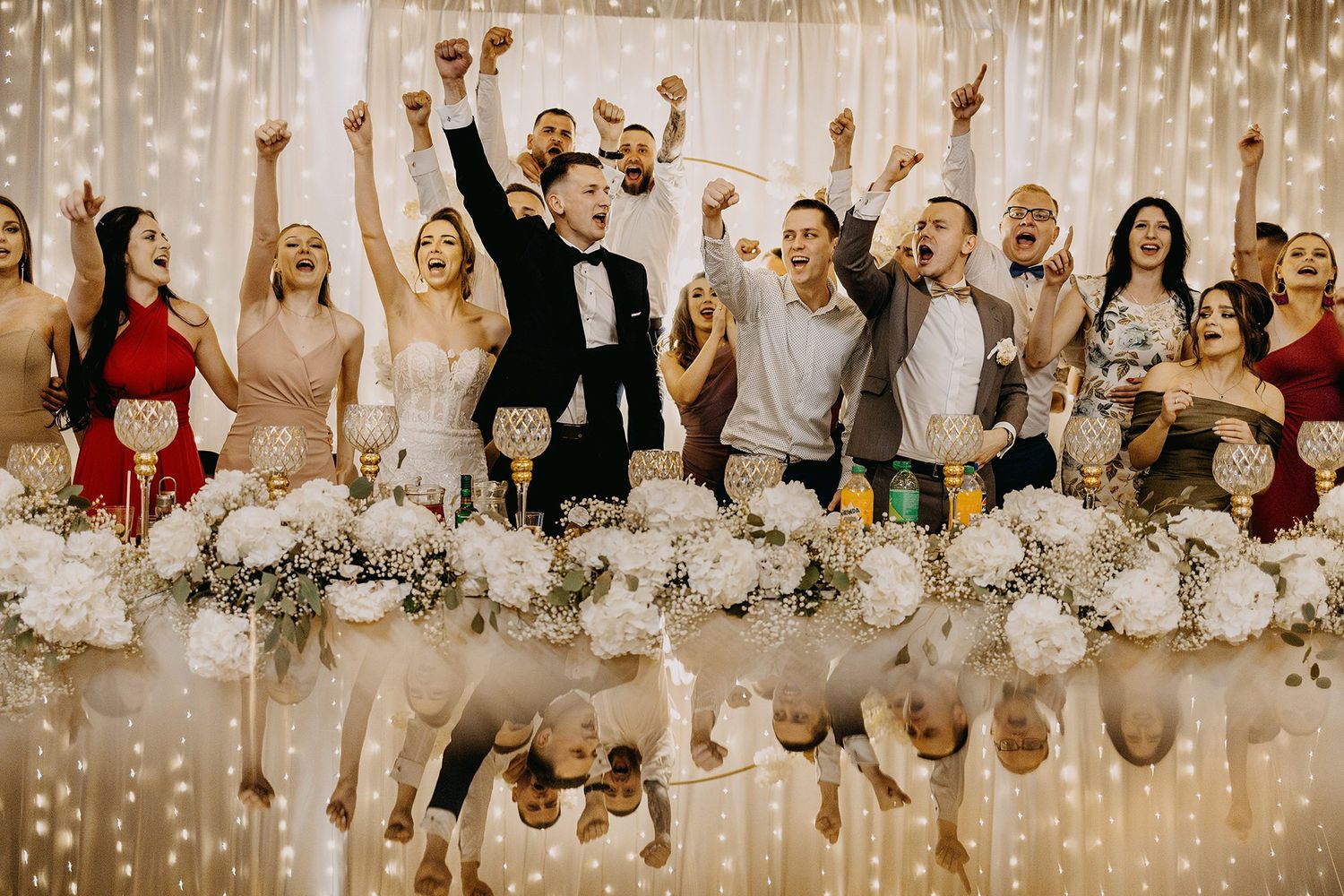 Joyful wedding party celebrating at reception table with floral arrangements and twinkling fairy lights backdrop.