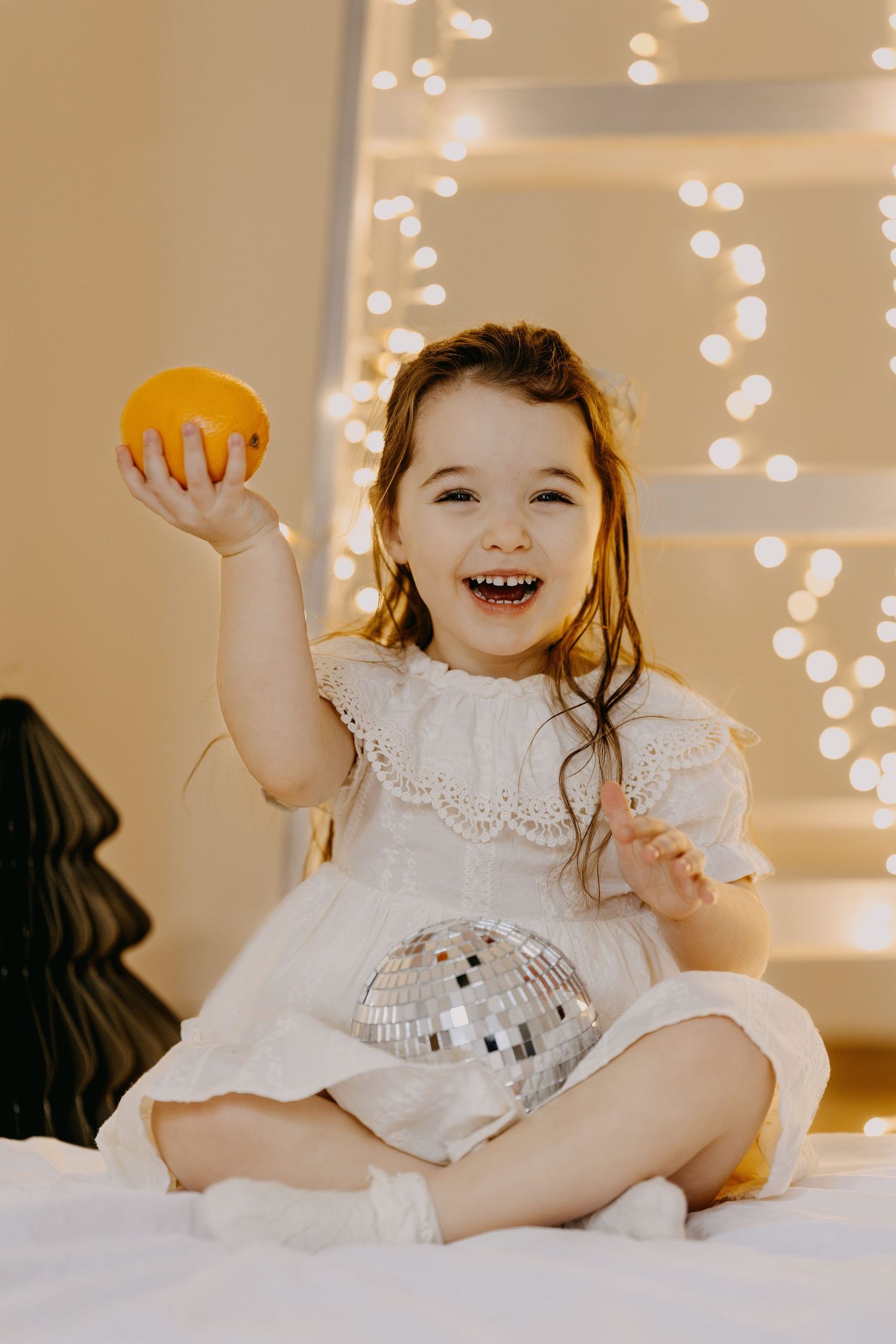 Smiling little girl in a white lace dress sits on a bed holding an orange and a disco ball, with warm string lights behind her.