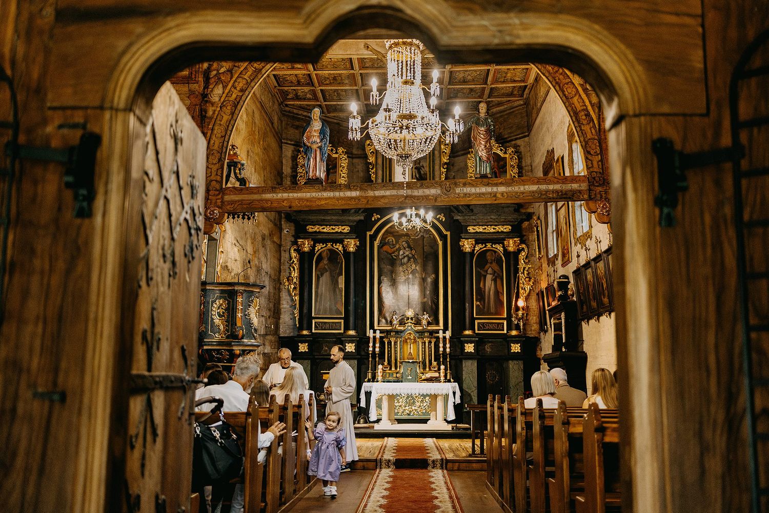 Interior of a historic wooden church with ornate black altars, chandelier, and clergy during a religious ceremony.