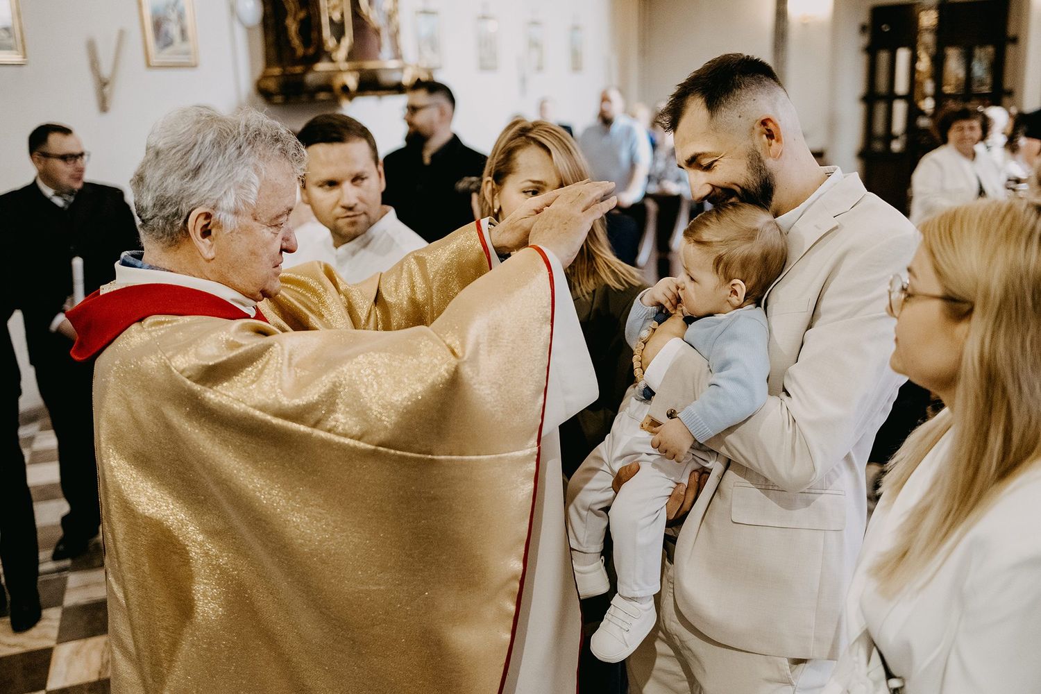 A priest performs a baptism blessing on a baby held by the father in a decorated church.
