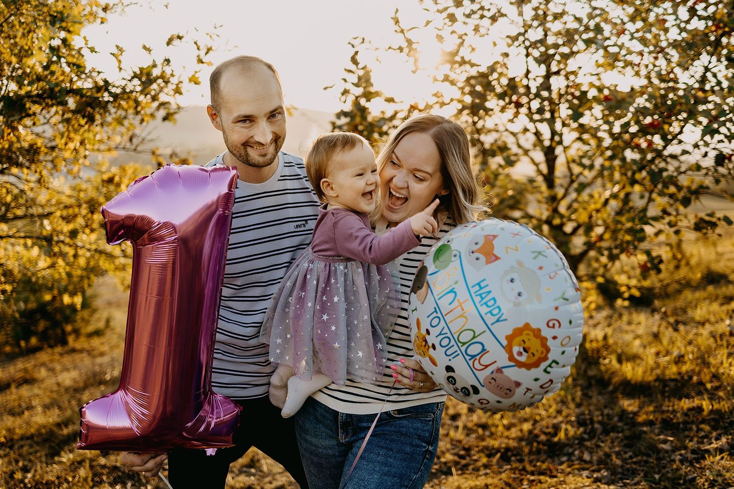 Family celebrating baby's first birthday with pink number one balloon outdoors.