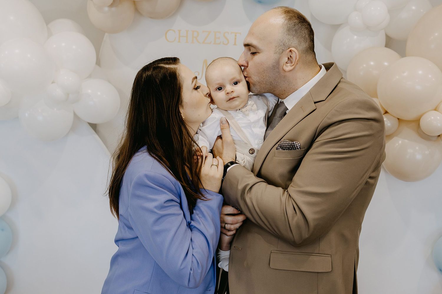 Parents kissing their baby on the cheeks at a christening party with balloon decorations.