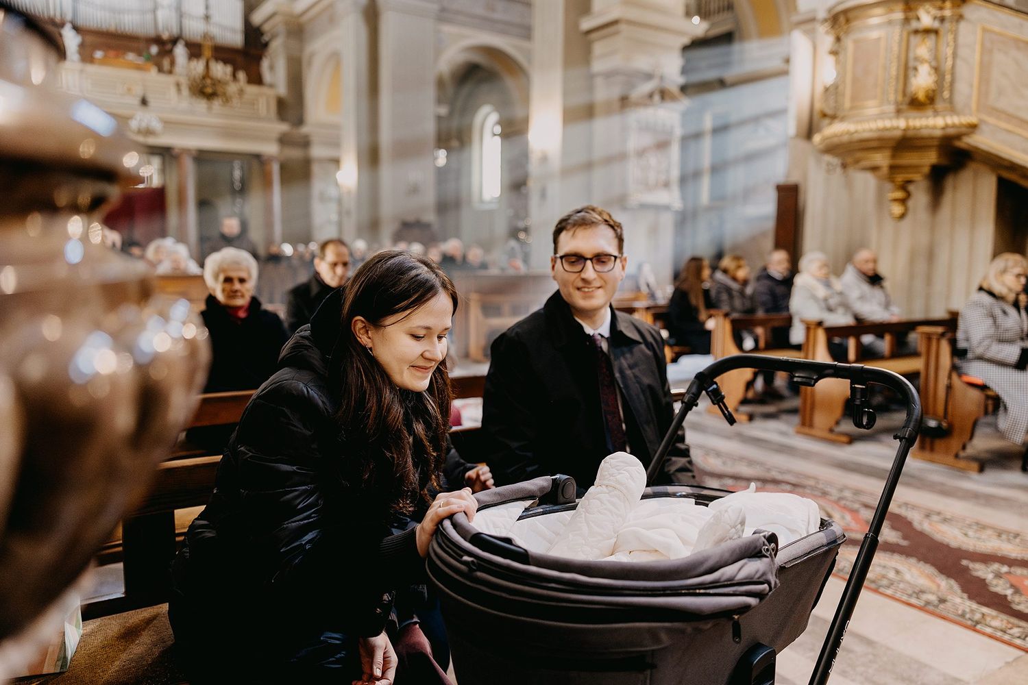 Parents with baby in stroller inside ornate church during baptism ceremony.