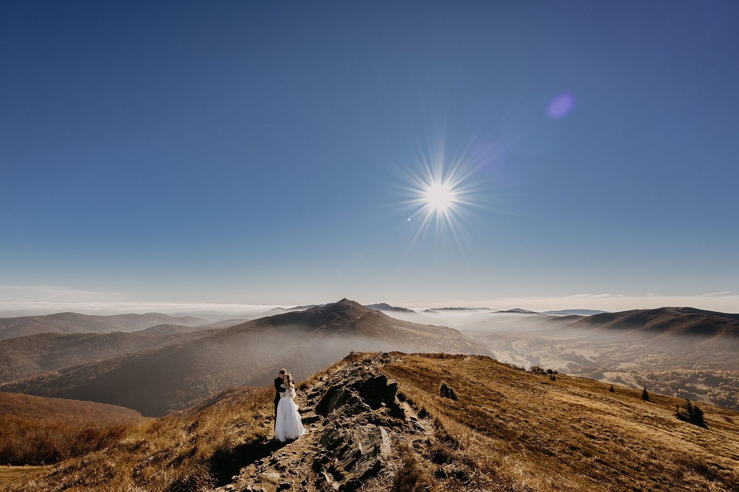 A bride and groom stand on a rocky mountain summit with stunning misty valley views and bright sunlight.