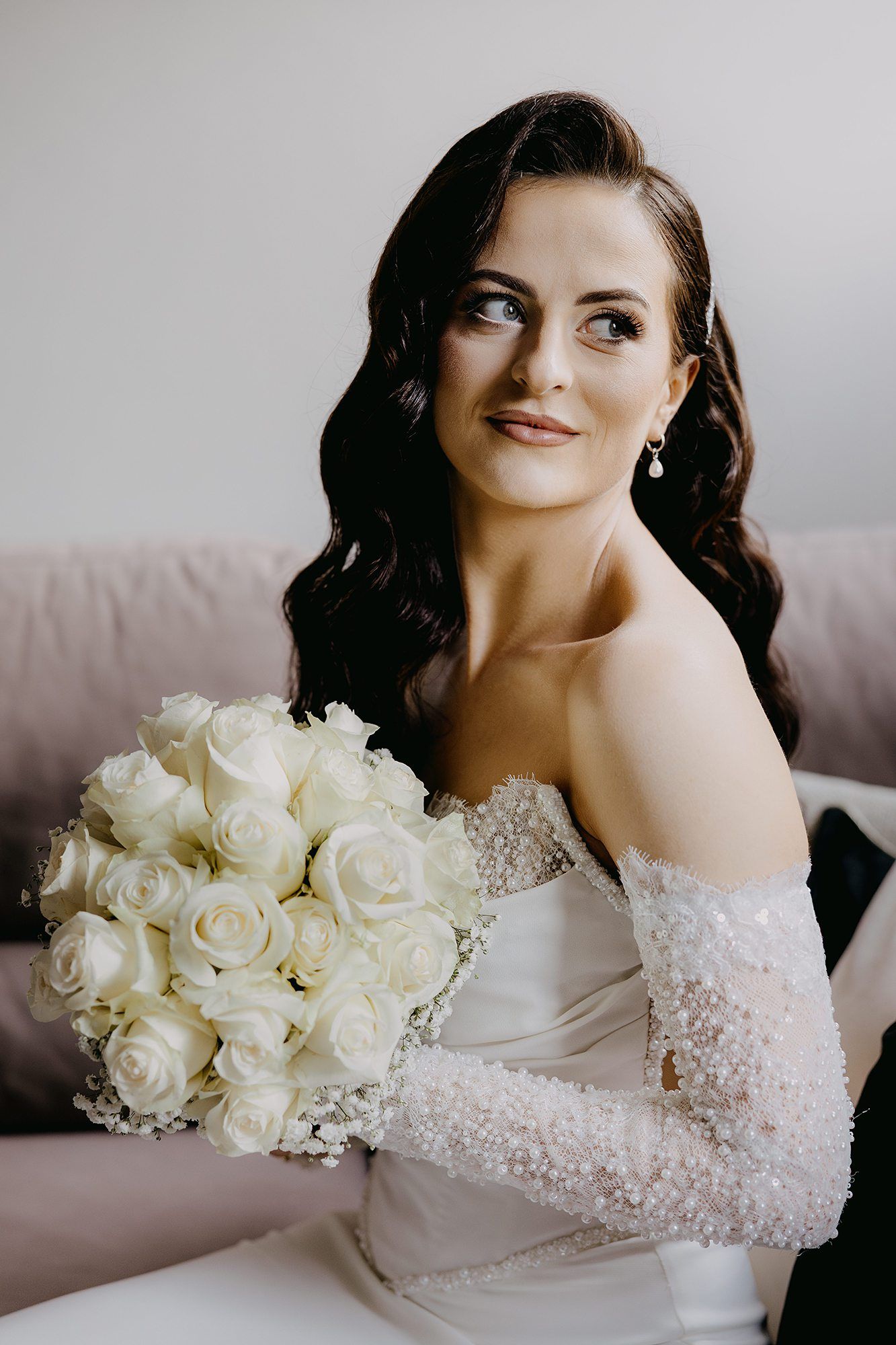 A beautiful bride with dark wavy hair holds a lush bouquet of white roses in a beaded off-shoulder gown.