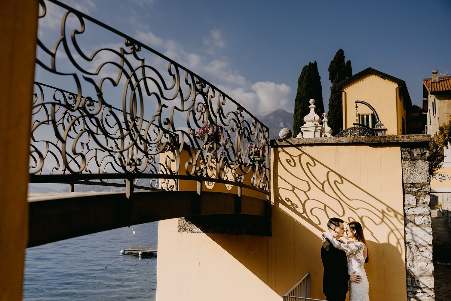 Wedding couple shares romantic kiss beneath ornate wrought iron bridge overlooking Lake Como in Italy.