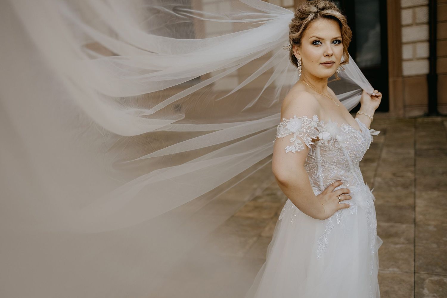 Bride in off-shoulder floral wedding gown poses gracefully as her veil billows dramatically in the wind.