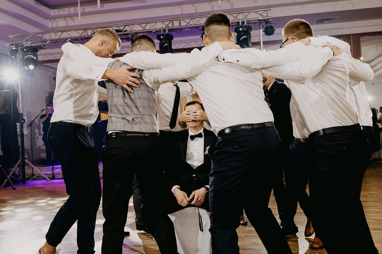 Groomsmen surround kneeling groom in playful dance floor celebration at wedding reception.