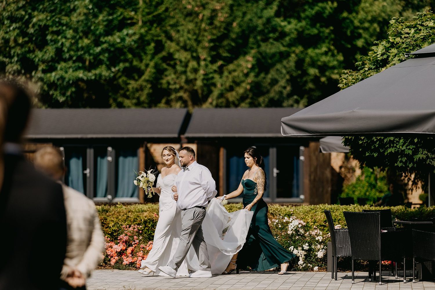 Bride in white gown walks with bridesmaid in emerald dress at outdoor wedding venue.