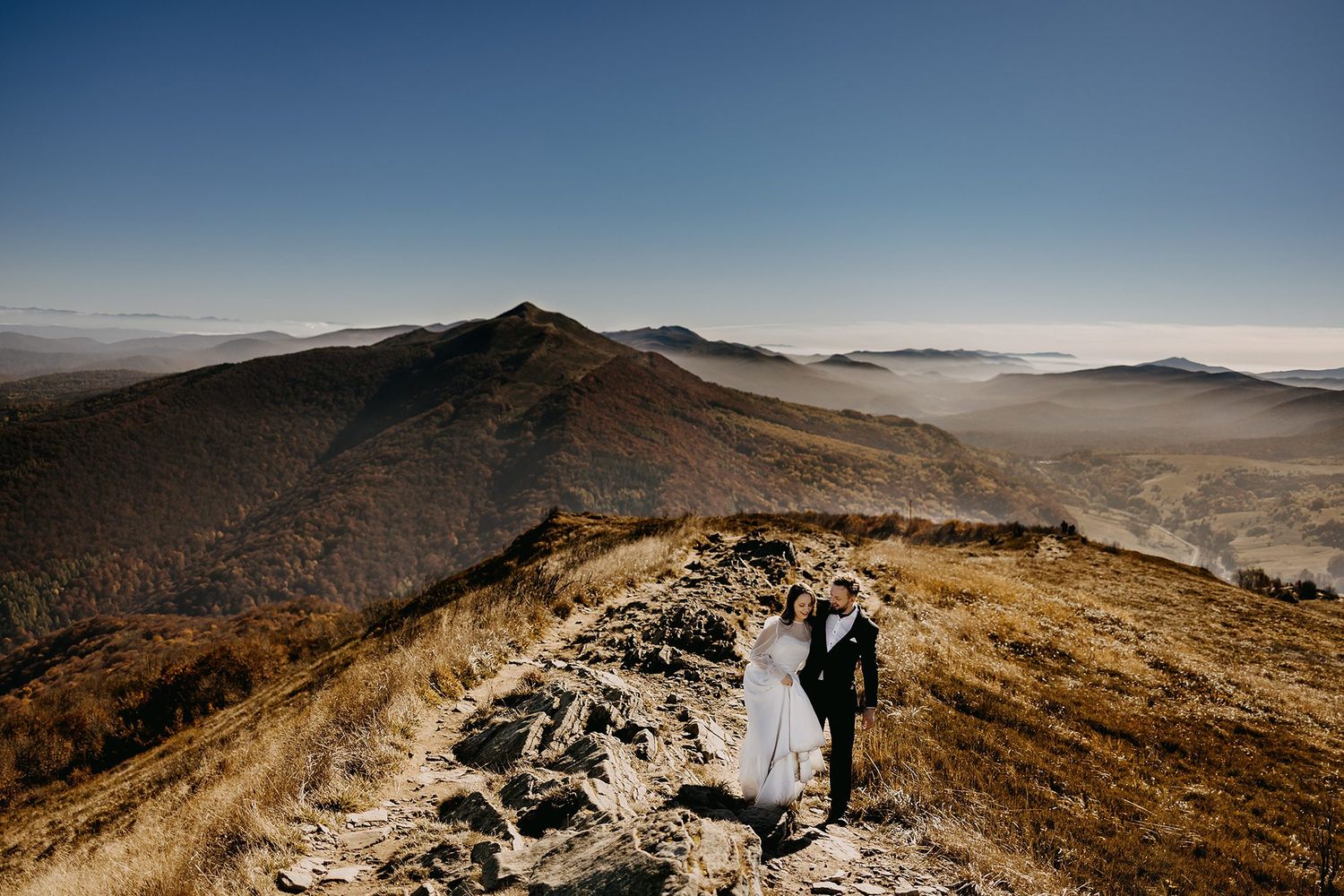 Wedding couple holding hands walking along a scenic mountain ridge under a clear blue sky.