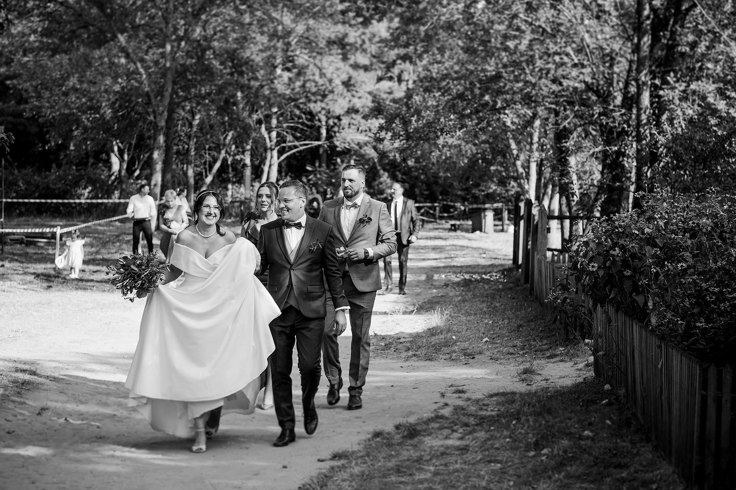 Black and white photo of wedding party walking along tree-lined path.
