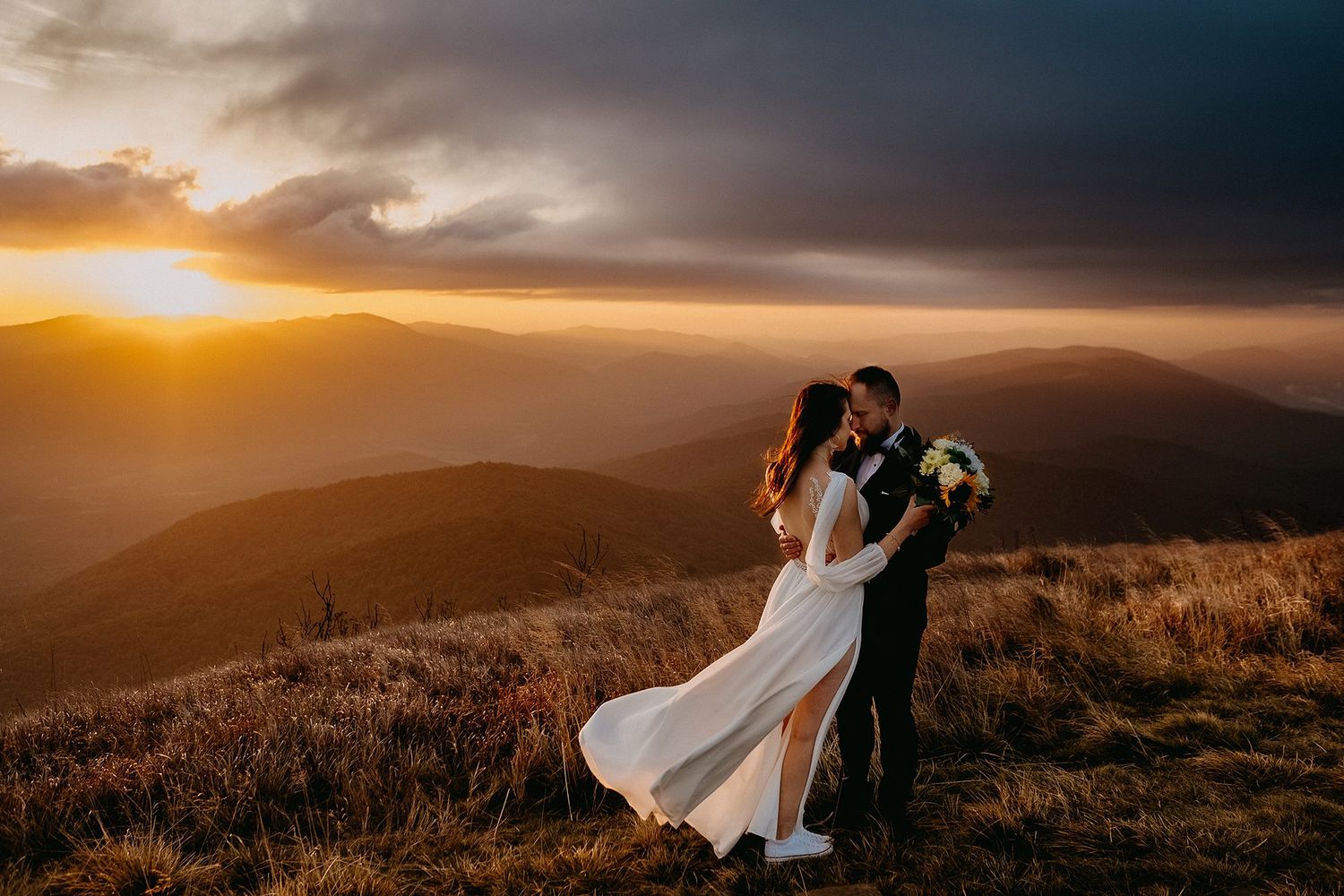 Newlyweds kissing on a mountain summit at dramatic golden sunset with rolling hills.