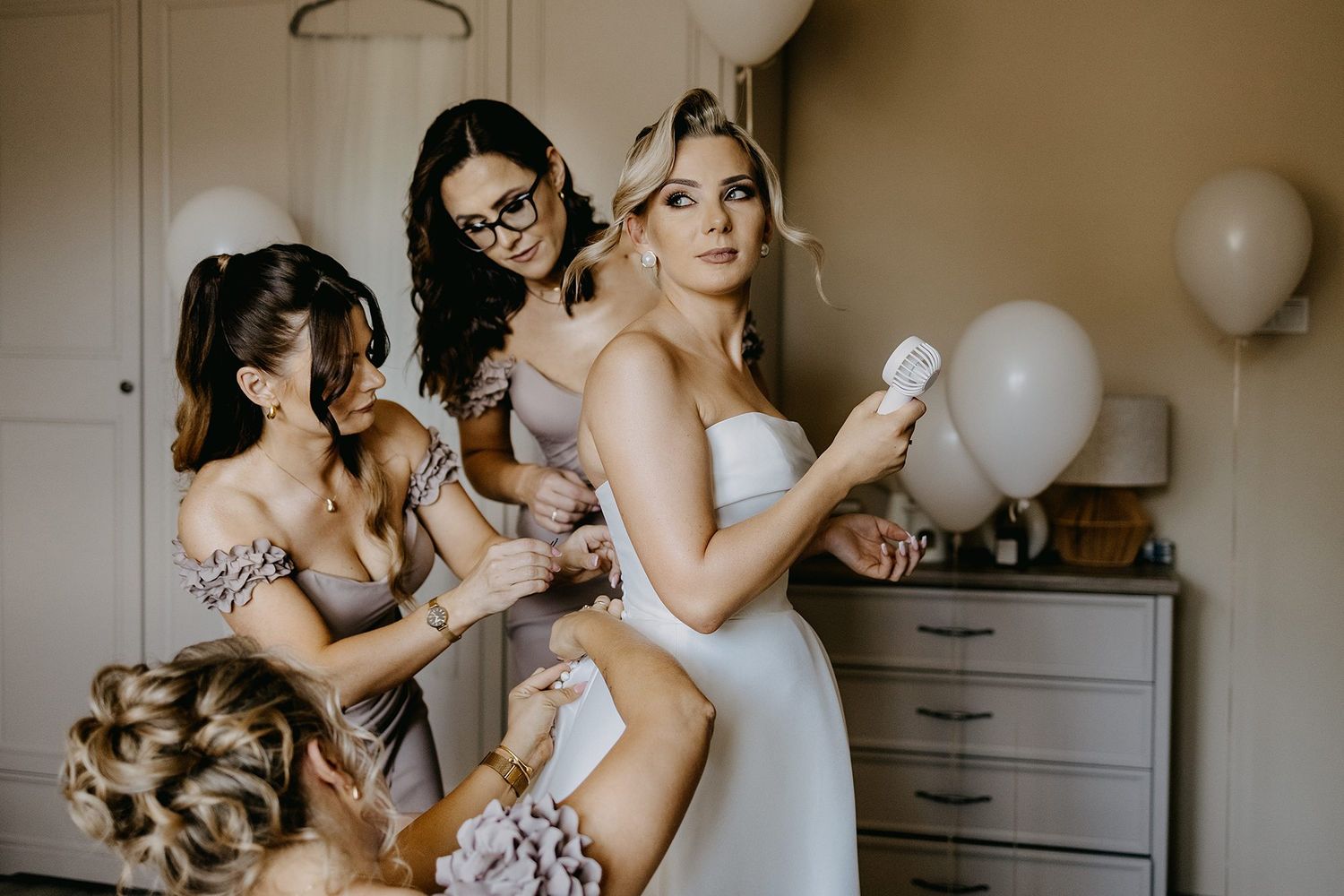 Bride in white strapless gown surrounded by bridesmaids in taupe dresses getting ready with balloons.