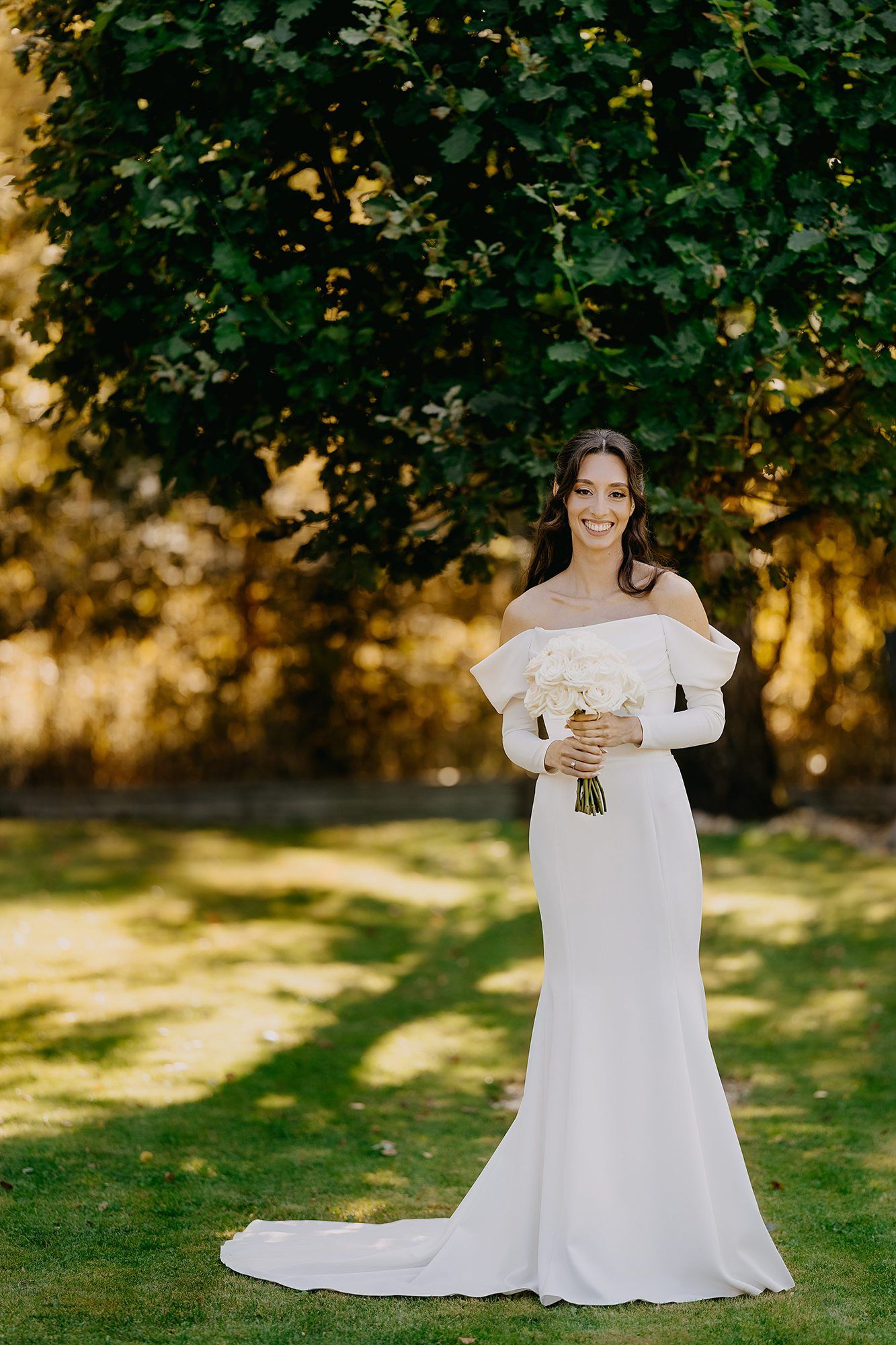 Elegant bride in off-shoulder white gown holding bouquet standing beneath a lush green tree outdoors.