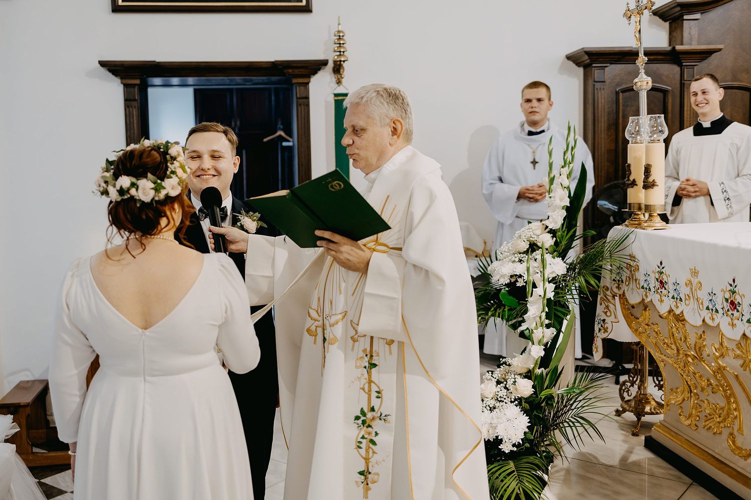 Bride and groom exchanging vows at church altar with priest and altar servers during wedding ceremony.