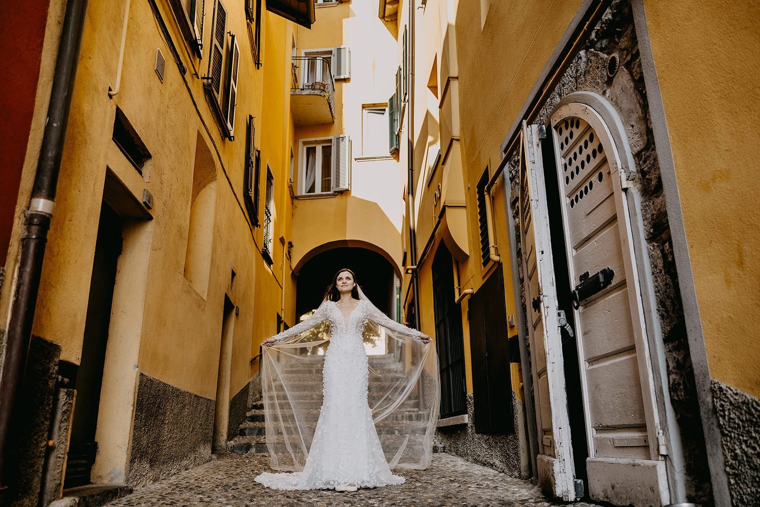 Bride in fitted white gown holds flowing veil wide open in a colorful Italian cobblestone alley.