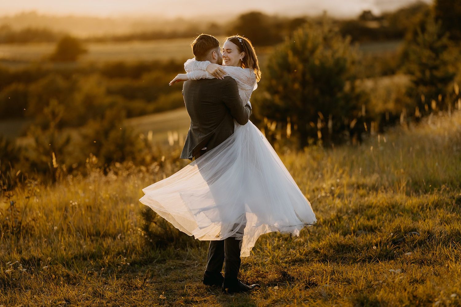 Groom joyfully lifting bride in flowing white dress during golden hour in a scenic meadow.