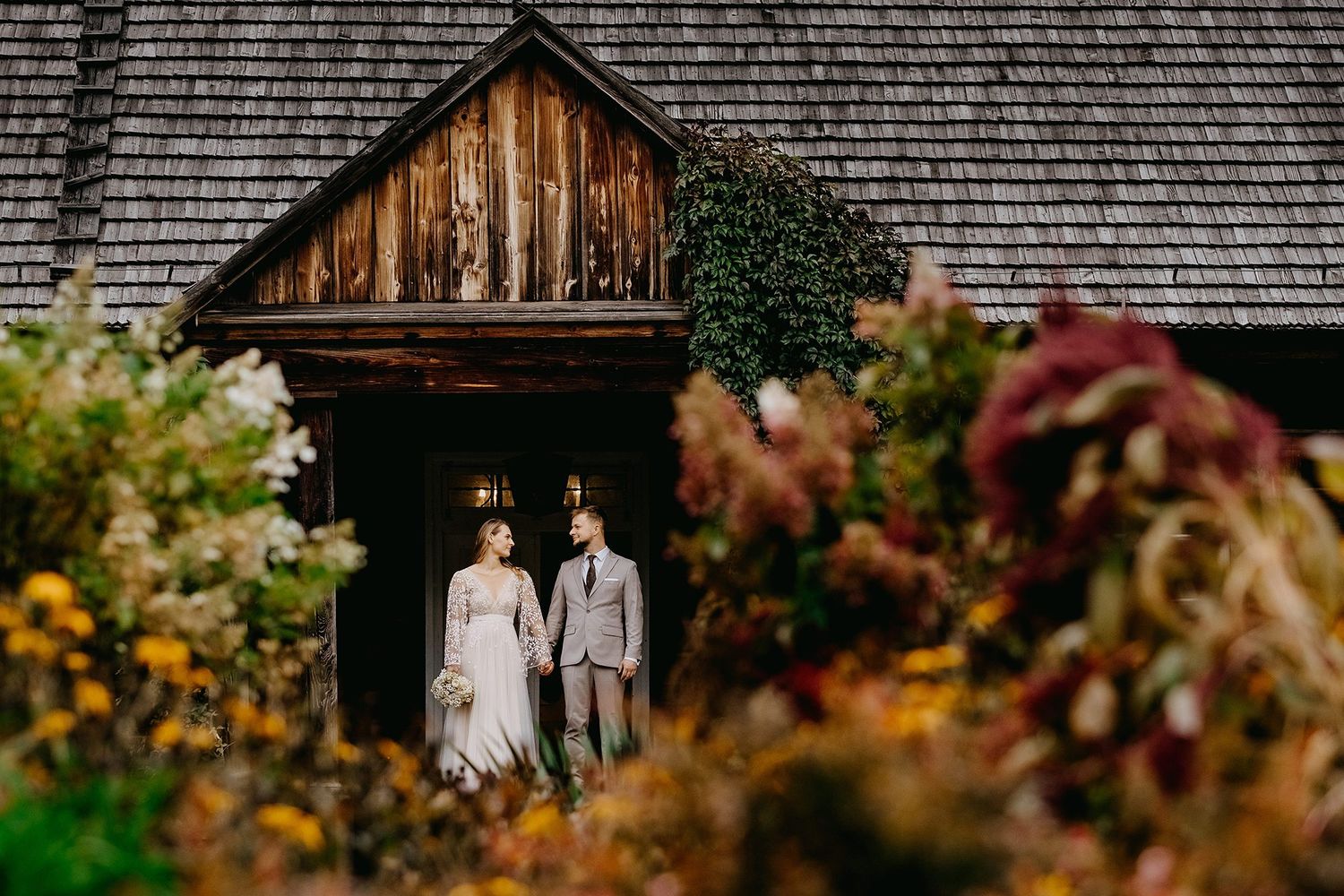 Bride and groom standing in a doorway of a rustic wooden building surrounded by colorful autumn flowers.