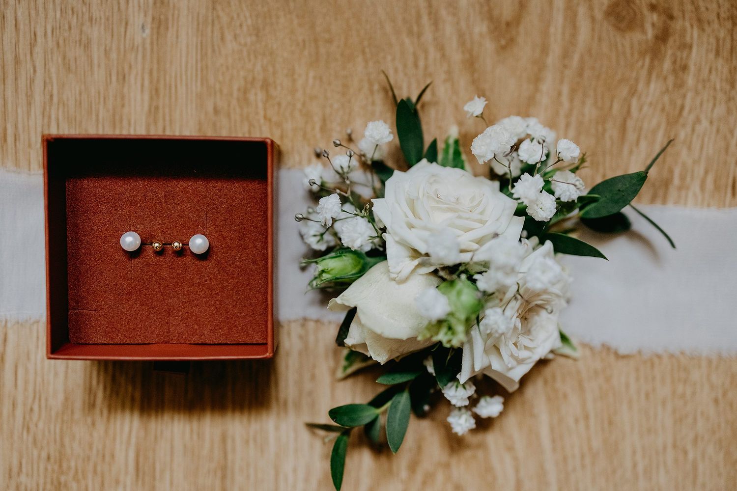 Pearl earrings in red jewelry box beside white rose boutonniere on wooden surface.