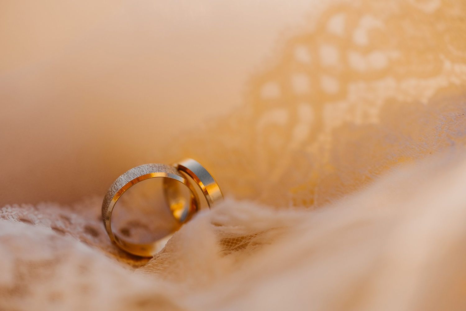 Two gold wedding rings rest intimately together on soft ivory lace fabric in warm light.