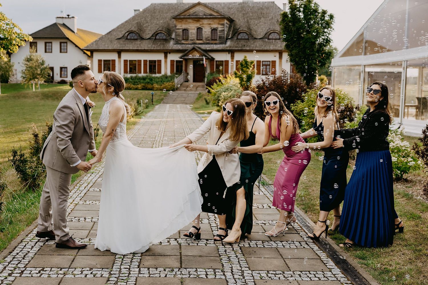 Bride in white gown laughing with bridesmaids in colorful dresses outside elegant manor house.