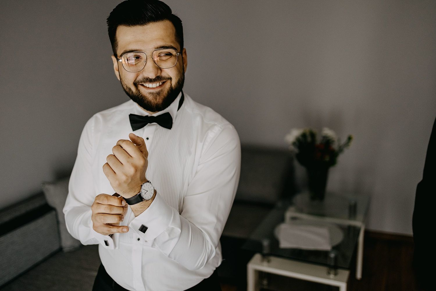 Smiling groom in white tuxedo shirt and bow tie adjusting his cufflinks while getting ready for wedding.