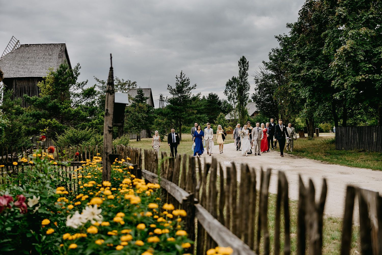 Wedding guests walk along a garden path lined with yellow flowers at a rustic outdoor venue.