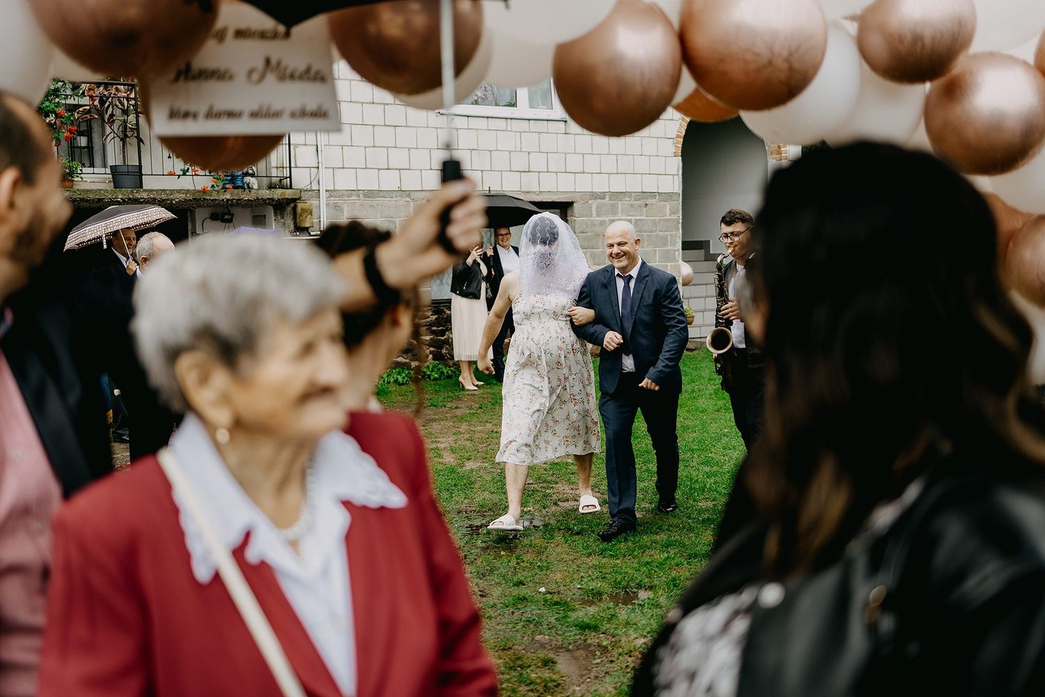 Bride in white veil walks with escort past rose gold balloons at an outdoor wedding celebration.
