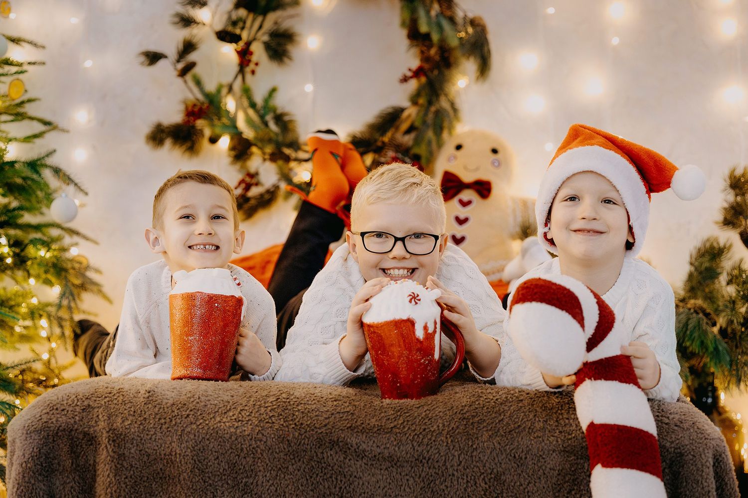 Three cheerful boys in holiday attire hold red mugs while posing in front of a decorated Christmas tree.