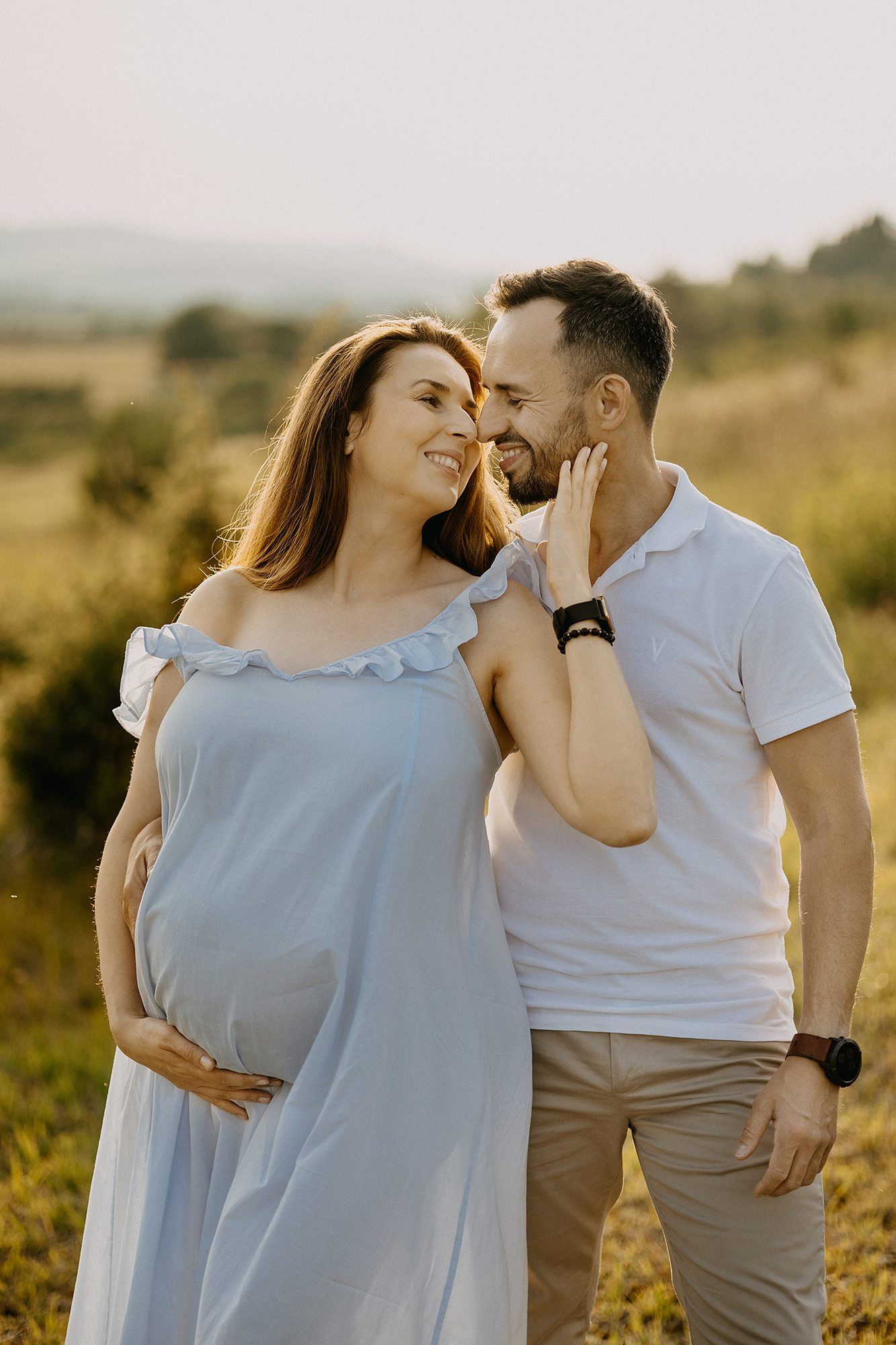 Happy pregnant woman in blue dress with partner in golden field at sunset, sharing a loving smile.