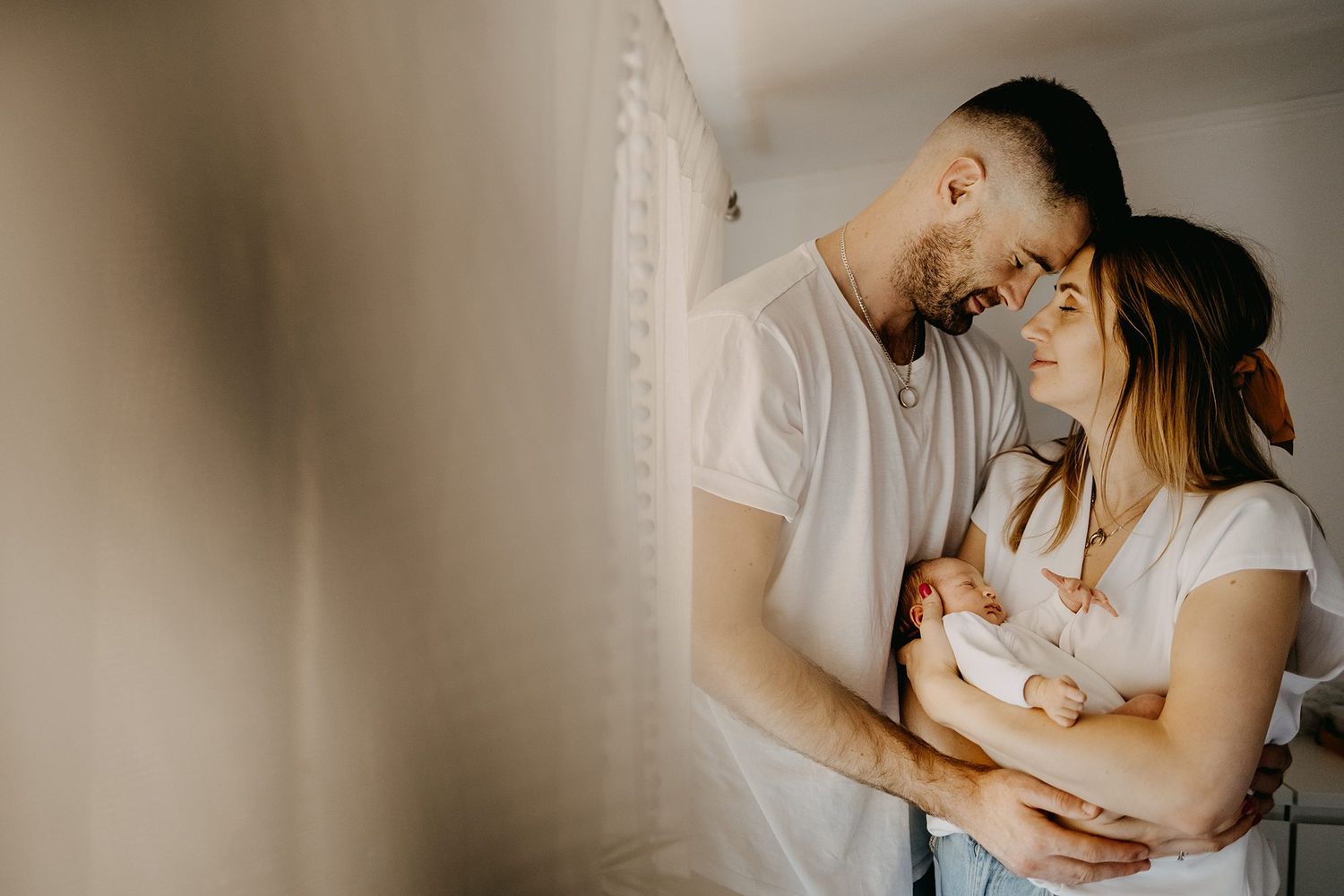 Happy parents in white shirts smiling over their newborn baby in a bright indoor setting.